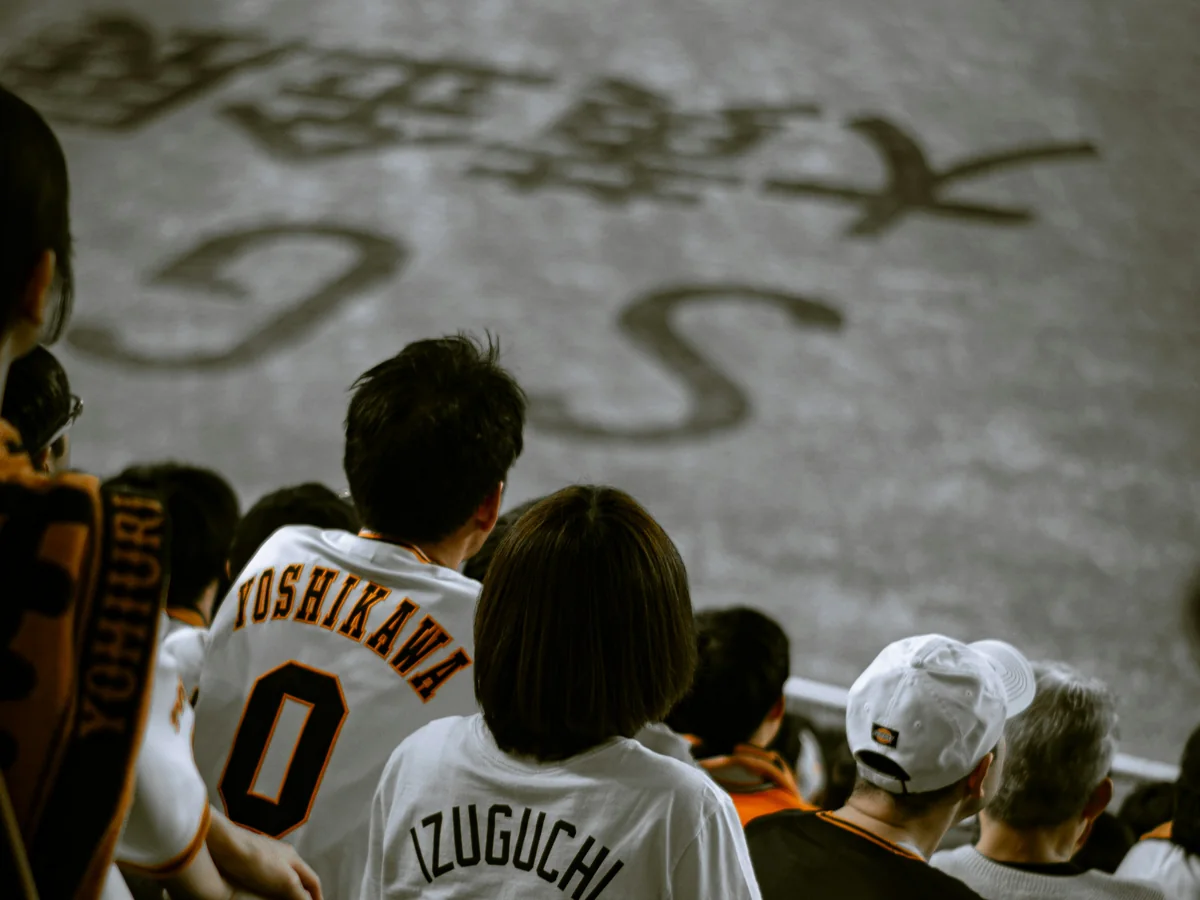Fans in white and orange Yomiuri Giants jerseys, seen from behind, watching the game from their seats
