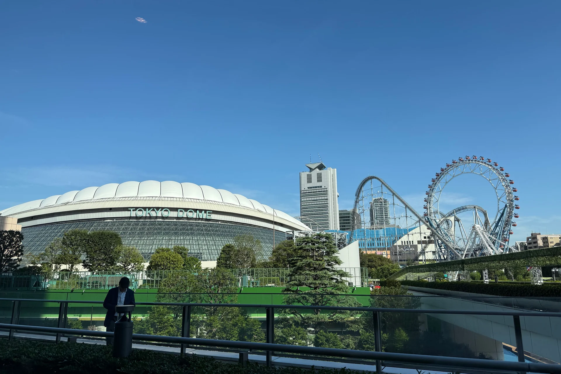 The white air-supported roof of Tokyo Dome with 'TOKYO DOME' lettering, photographed in daylight from outside the stadium, with the Tokyo Dome City amusement park visible behind it